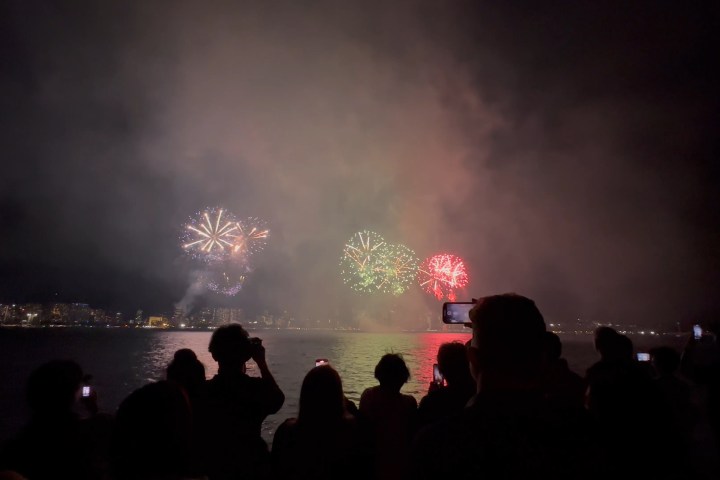 Silhouetted crowd watches colorful fireworks over water at night.