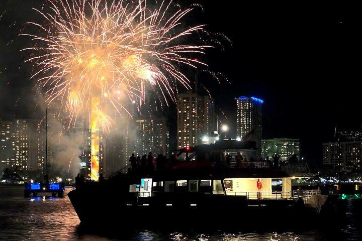 Nighttime fireworks display over a city skyline with a boat in the foreground.