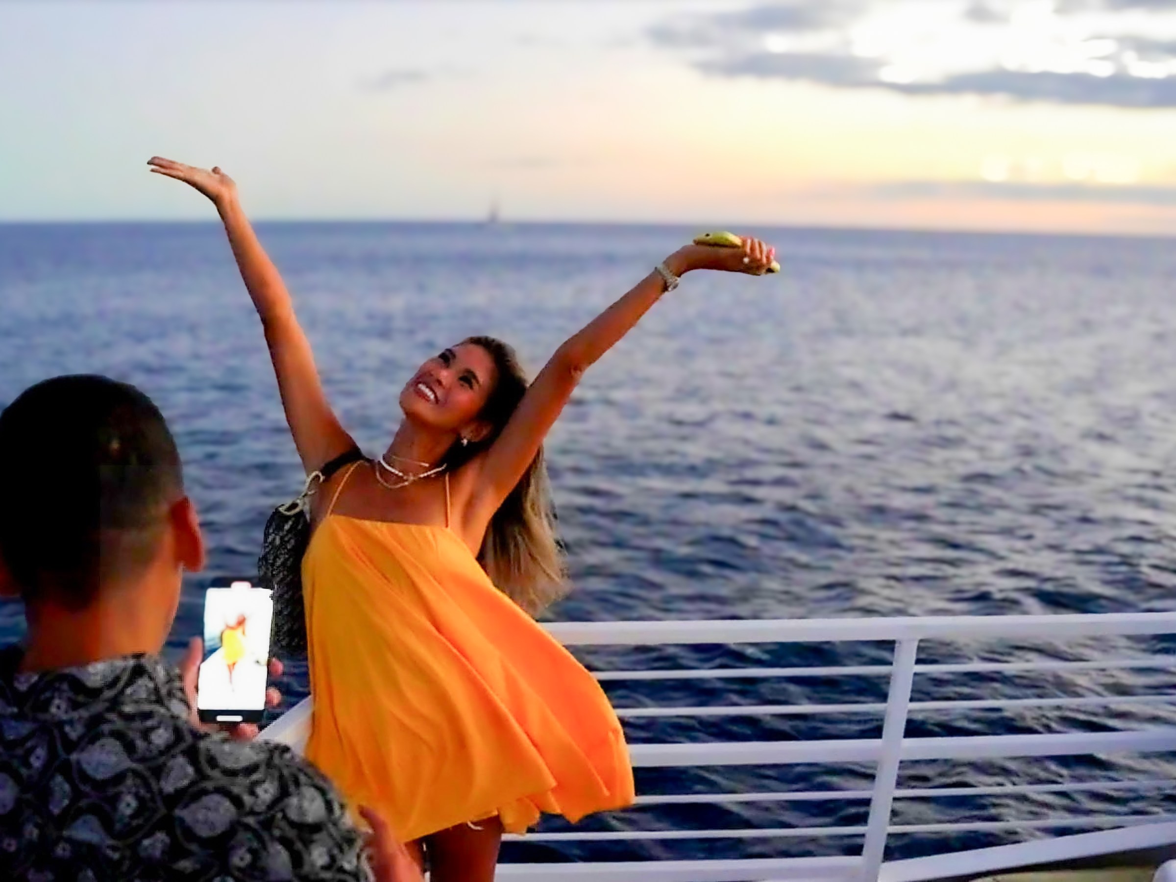 Woman poses happily on a boat at sunset, with a man photographing her.