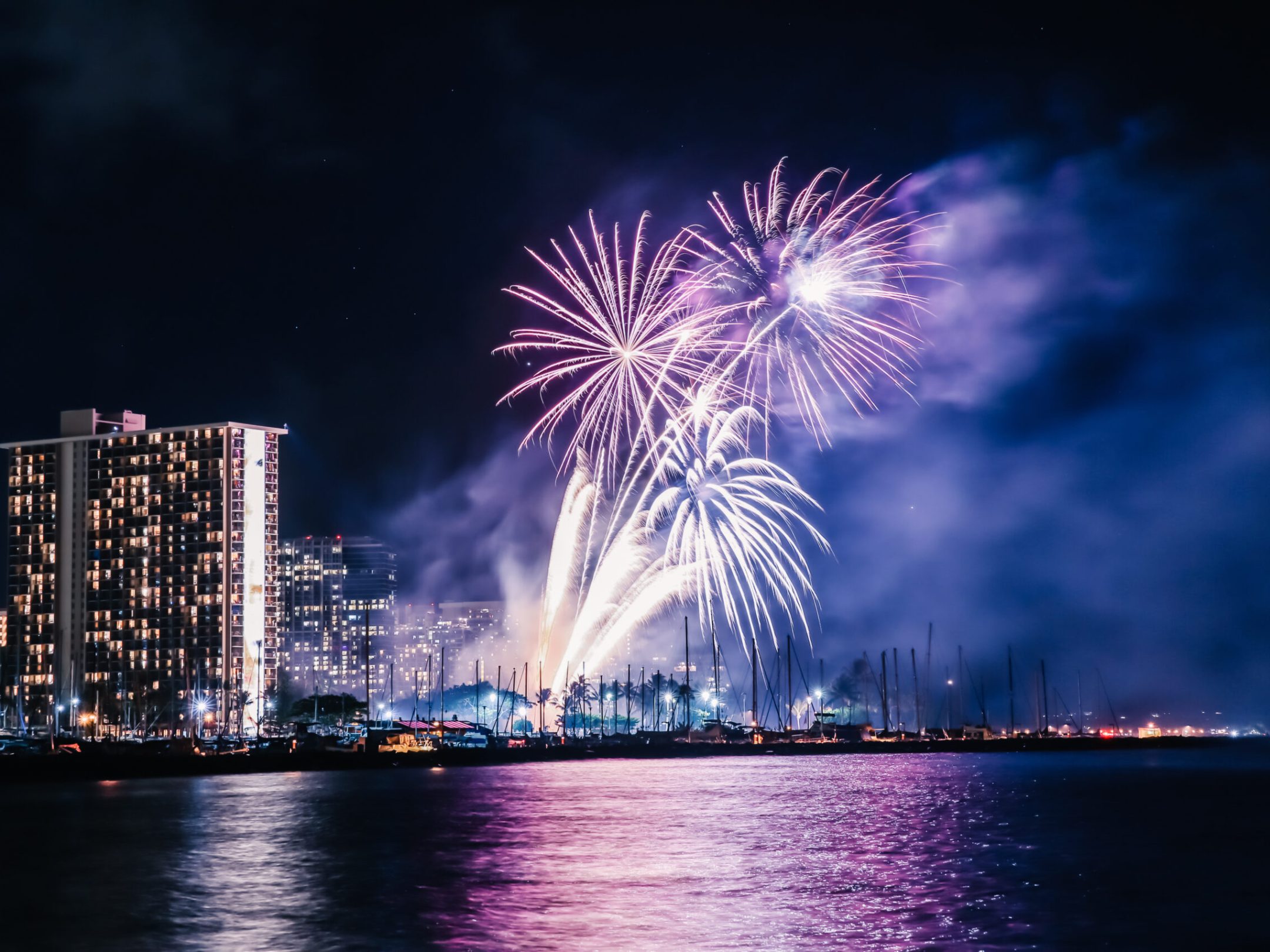 Fireworks over a city skyline by the water at night, with reflection on the water surface.