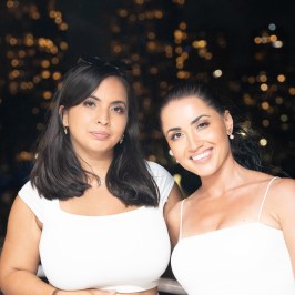 Two women in white tops smiling with a city nightscape in the background.