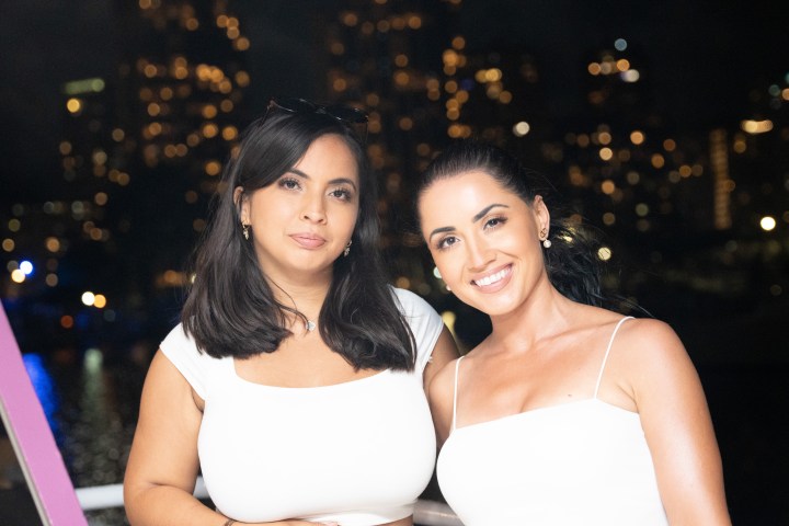 Two women in white tops smiling with a city nightscape in the background.