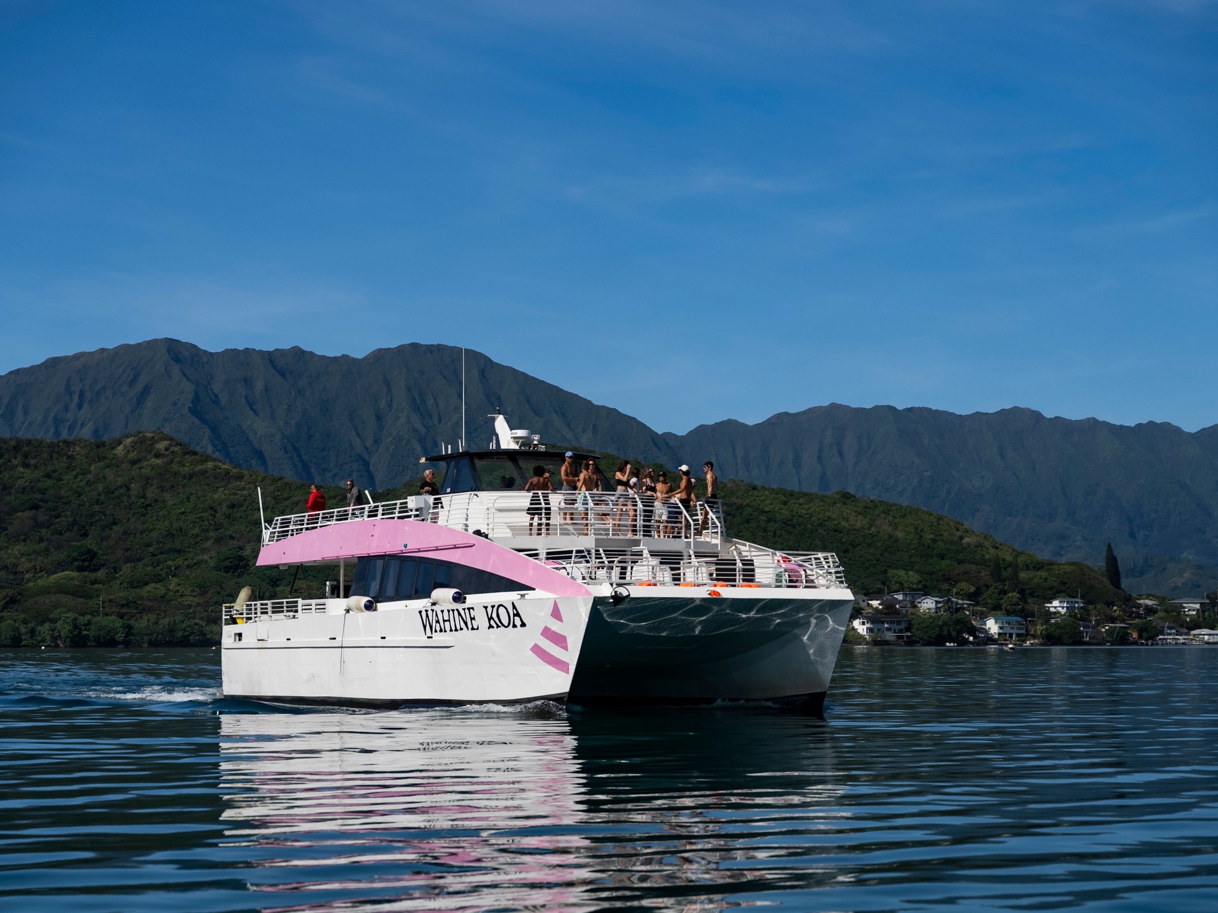 Catamaran on water with mountains in background, people on deck, clear blue sky.