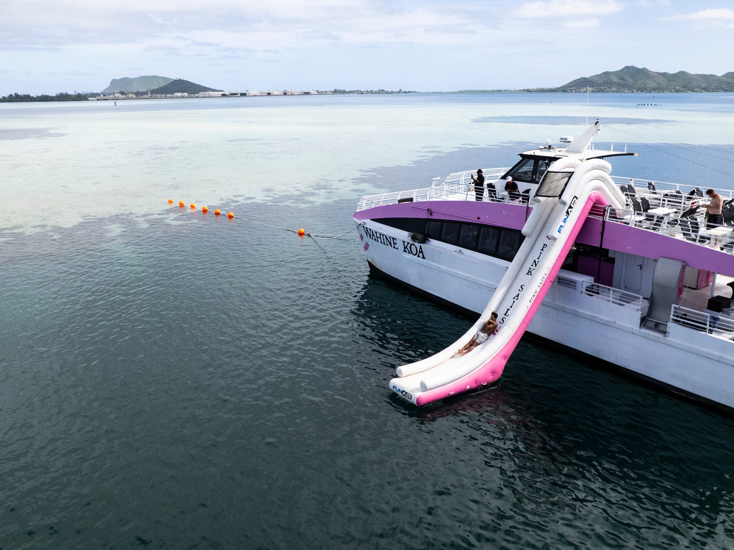 Boat with a pink slide docked on a calm blue sea, mountains in the background.