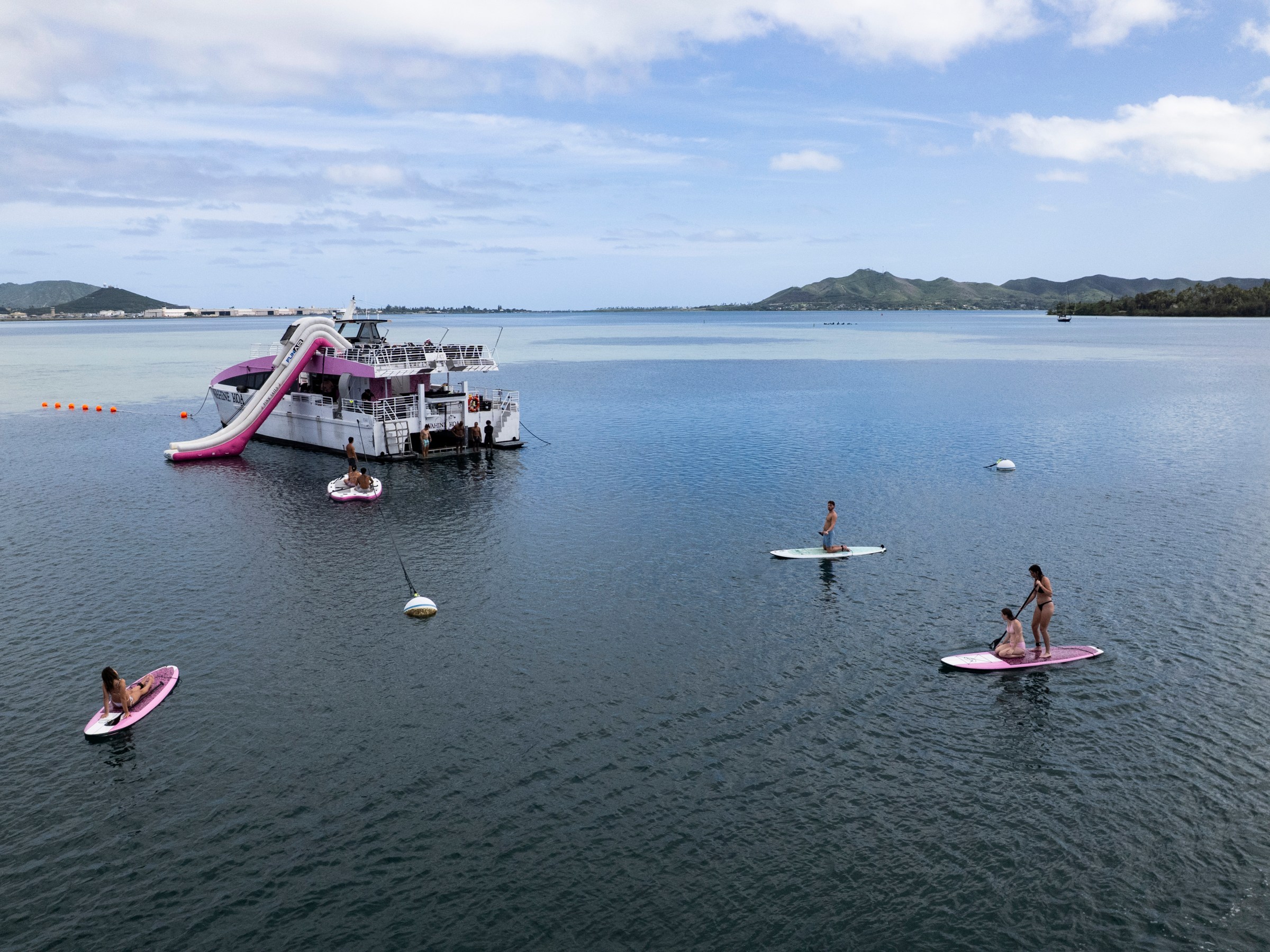 People paddleboarding near a boat with a slide in a scenic bay under a partly cloudy sky.