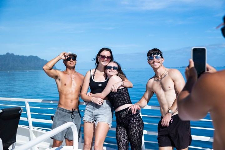 Four people posing on a boat with ocean and mountains in the background, one taking a photo with a smartphone.