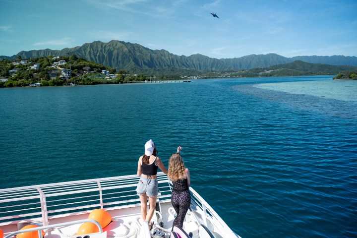 Two people on a boat admire the mountains and blue water on a sunny day.
