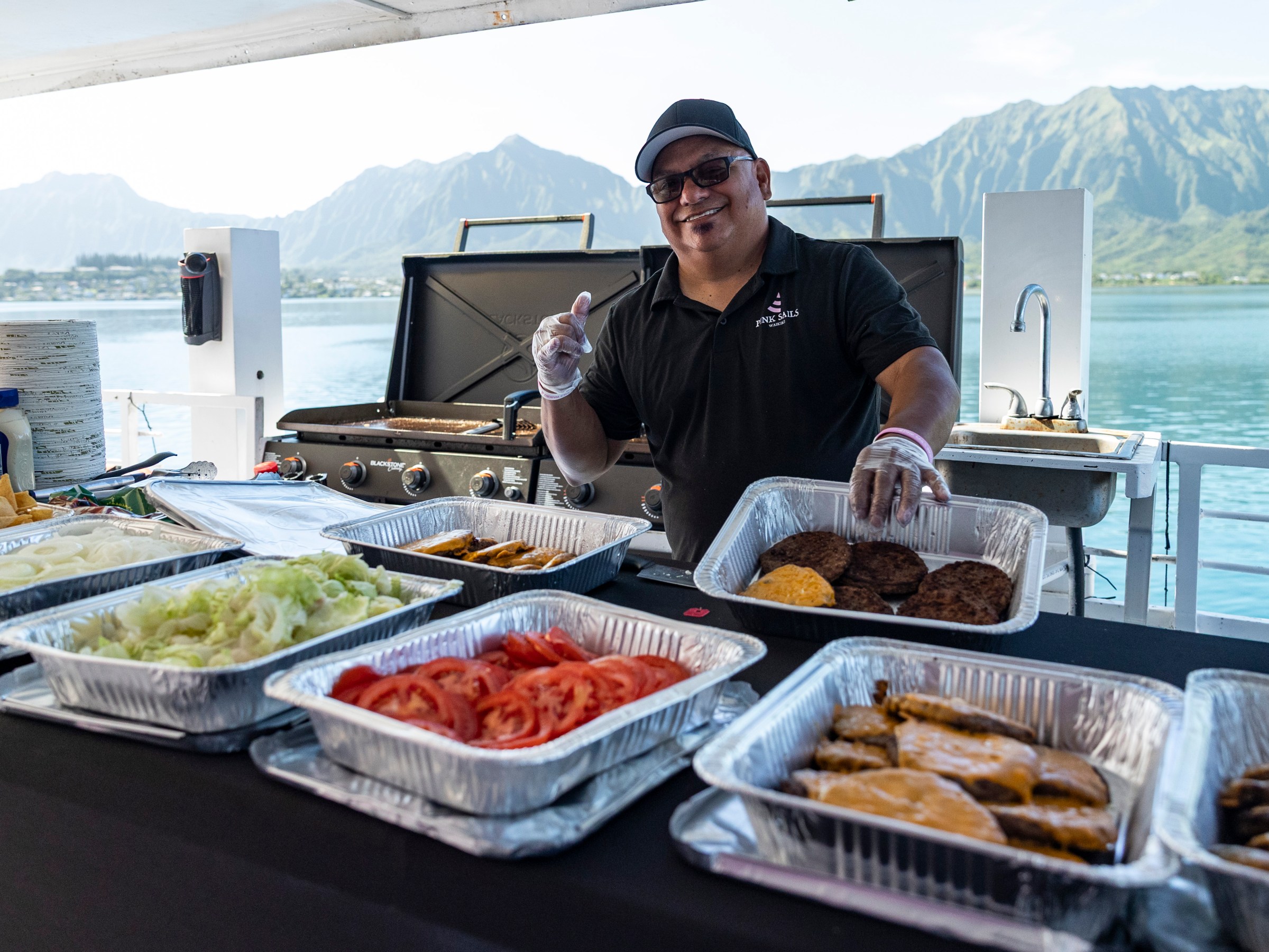Man on boat grilling burgers with trays of toppings, mountains in background.