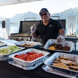 Man on boat grilling burgers with trays of toppings, mountains in background.