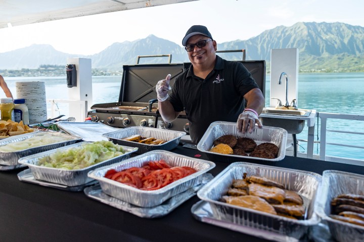 Man on boat grilling burgers with trays of toppings, mountains in background.