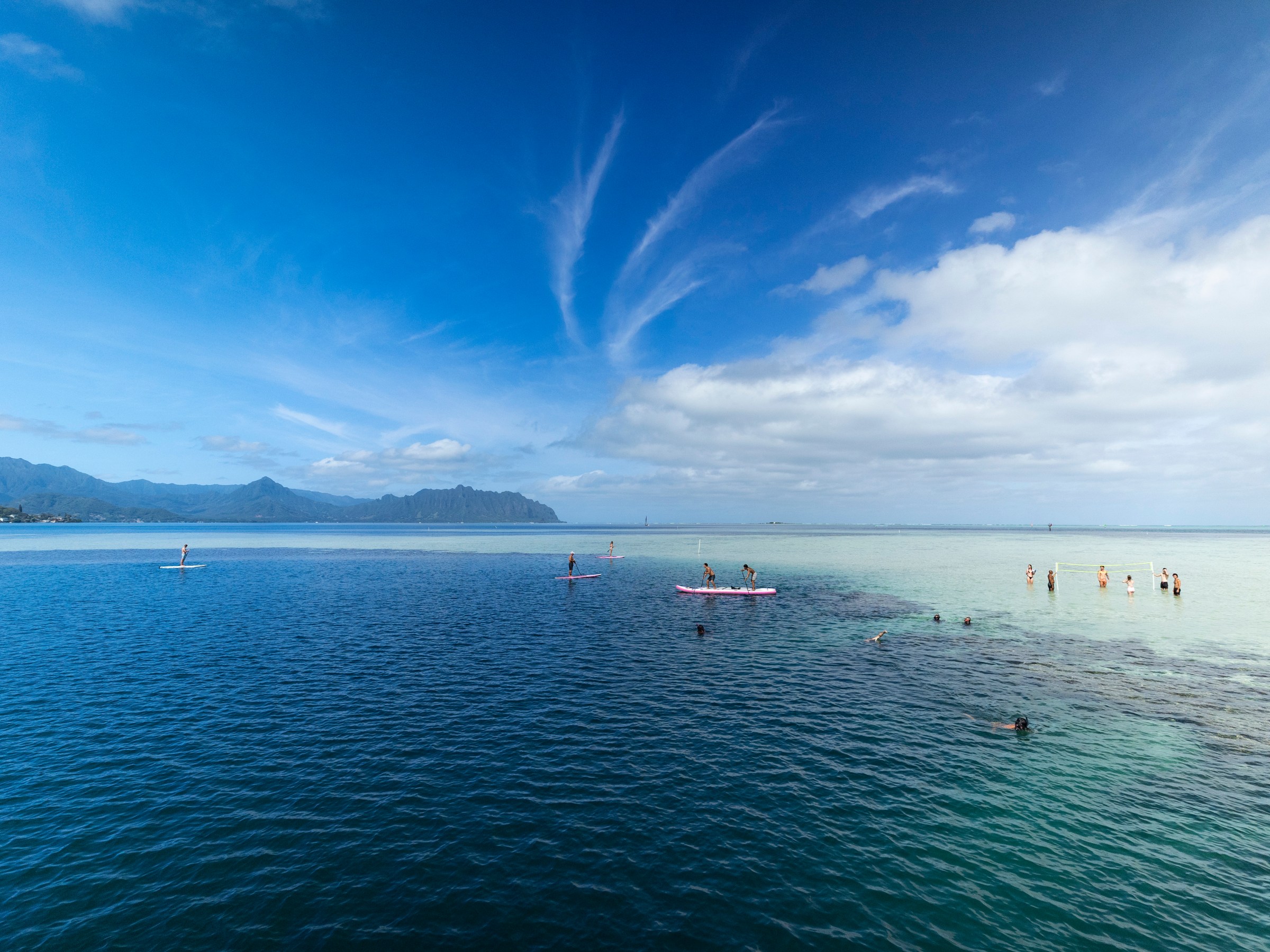 People paddleboarding and standing in clear blue ocean against a mountain backdrop.