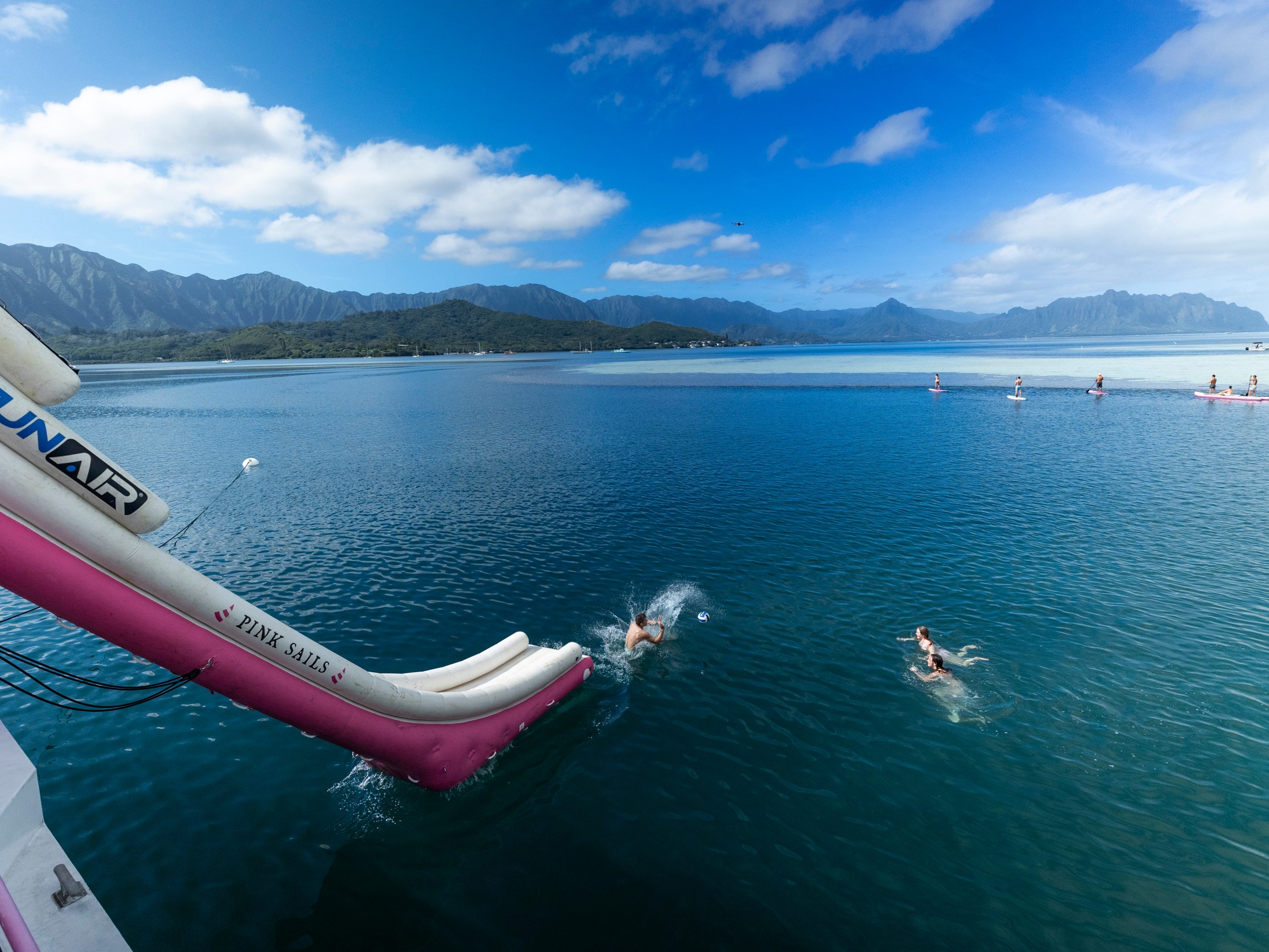 People swimming and paddleboarding near a slide into the ocean under a blue sky.