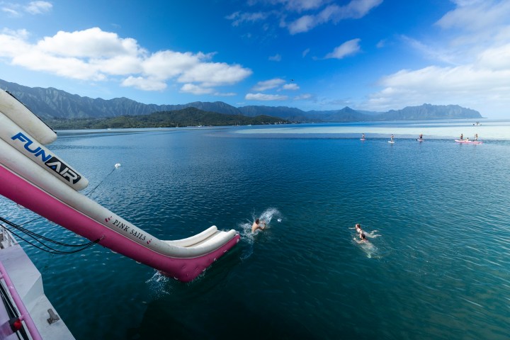 People swimming and paddleboarding near a slide into the ocean under a blue sky.