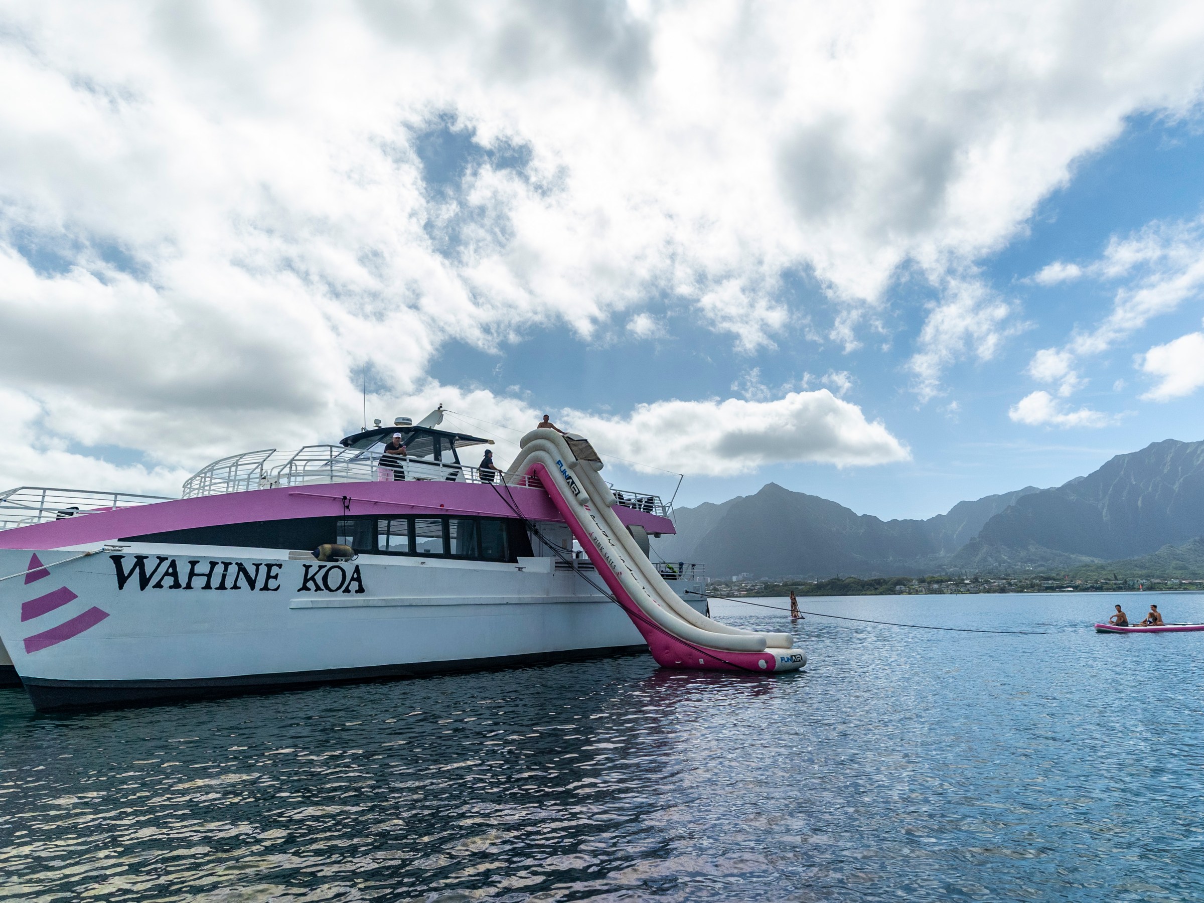 Boat named Wahine Koa with a slide on water, mountains in background, and two people on a kayak.