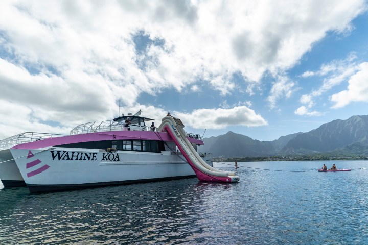 Boat named Wahine Koa with a slide on water, mountains in background, and two people on a kayak.
