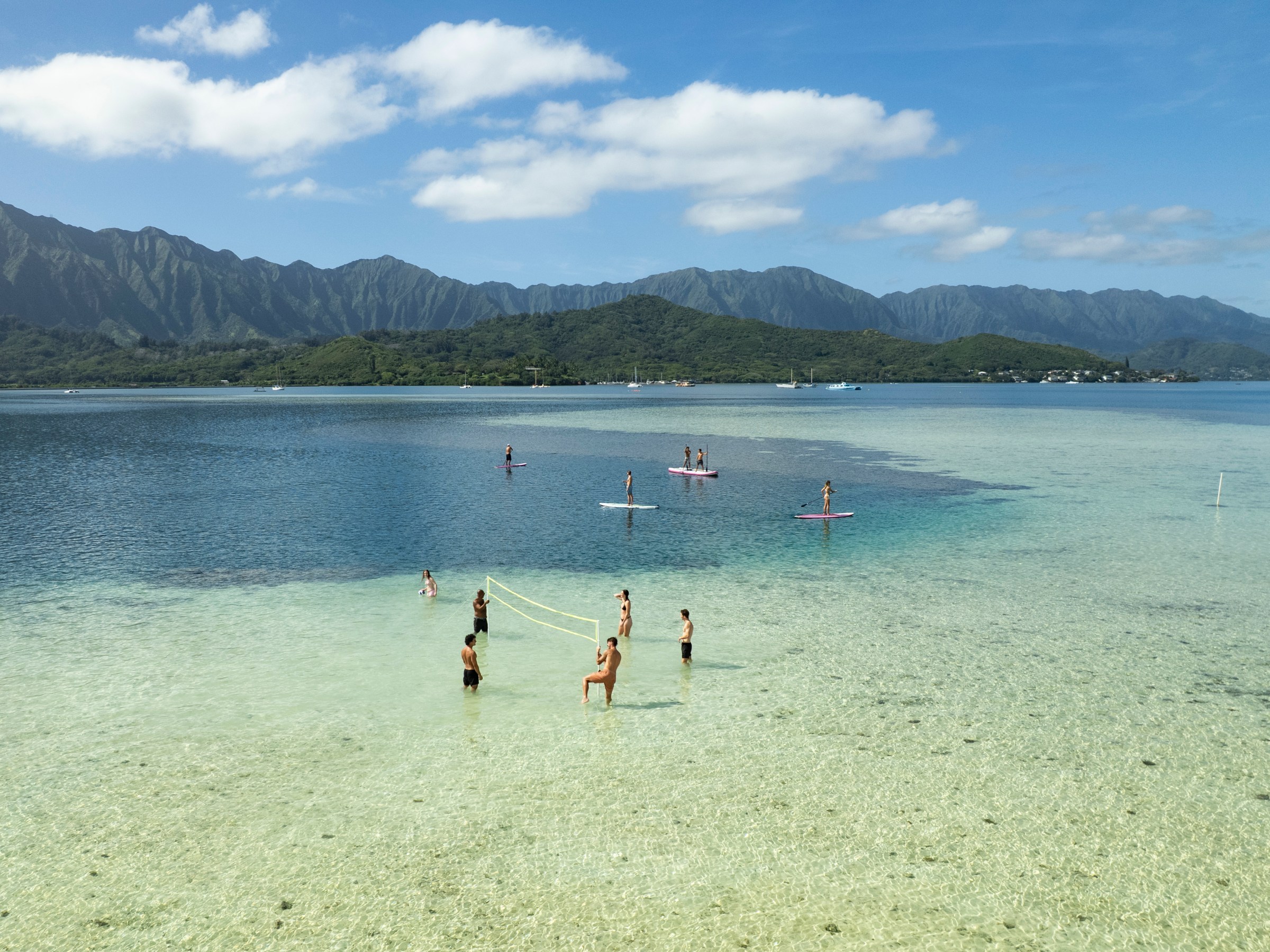 People in shallow ocean water, some playing volleyball, others paddleboarding, with mountains in the backdrop.