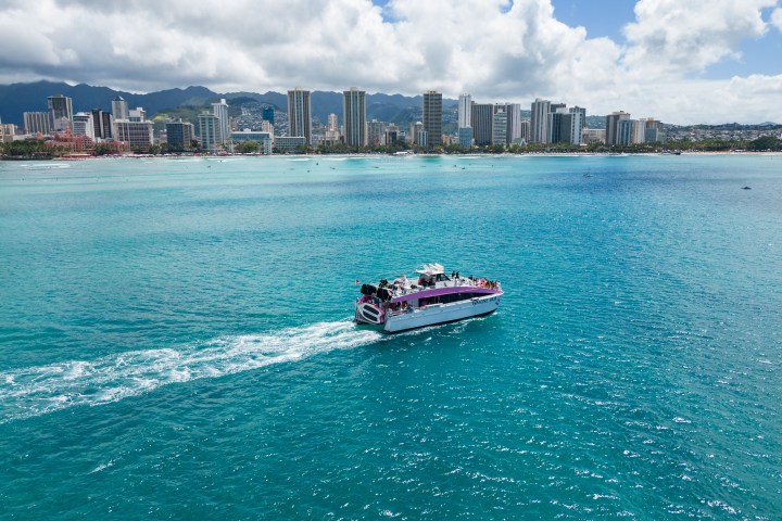 Boat on turquoise water with city skyline and mountains in the background.