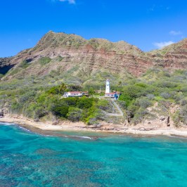 Coastal landscape with a lighthouse near turquoise water and a mountain backdrop.