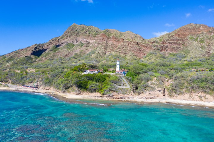Coastal landscape with a lighthouse near turquoise water and a mountain backdrop.