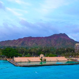 Coastal view of a mountain with buildings and blue ocean in the foreground at sunset.